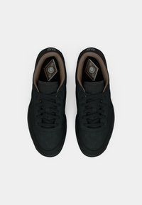 Melna Reebok sporta apavu pāris ar brūnu iekšējo oderi, melnām auklām un apzīmētām iekšējām zolēm, skatīts no augšpuses uz balta fona.