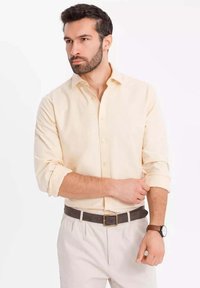 Helles gelbes Button-up-Hemd mit aufgerollten Ärmeln, kombiniert mit hellen Hosen und einem braunen Gürtel. Ein Armbanduhren wird am linken Handgelenk getragen.