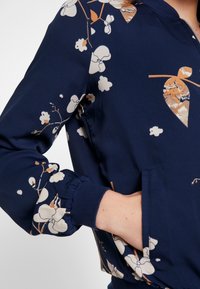 Námořnický modrý květinový kabát z lehké látky, s elastickými manžetami a dvěma bočními kapsami; zdobí ho krémové a rezavé květinové vzory.