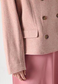 Roosa tweed topeltkootud jakk pruunide nuppudega, laiad varrukad ja küljetaskud, koos roosa seelikuga. Tekstureeritud kangas peene mustriga.