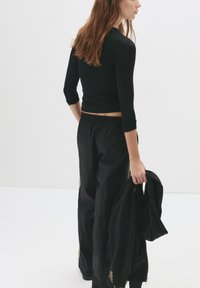 Schwarzes Langarmtop und lockere schwarze Hose mit elastischem Bund. Das Model hält eine passende schwarze Jacke und zeigt ein minimalistisches Design.
