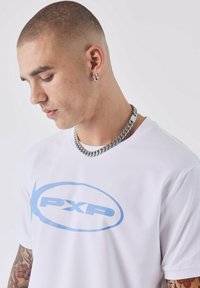 Camiseta blanca con el logo azul "PXP", cuello redondo, mangas cortas, textura suave. El modelo lleva un collar de cadena plateada y tiene tatuajes visibles.