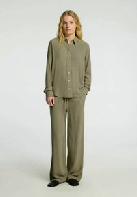 Vrouw die met handen in de zakken staat, gekleed in een losse olijfgroene overhemd met knopen en bijpassende brede broek, met zwarte schoenen.
