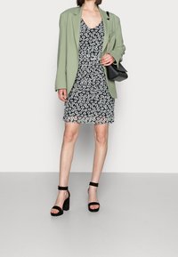 Grüner Blazer über einem floral gemusterten schwarzen Kleid mit kleinen weißen und blauen Blumen. Schwarze Plateaustiefel und eine kleine schwarze Handtasche.