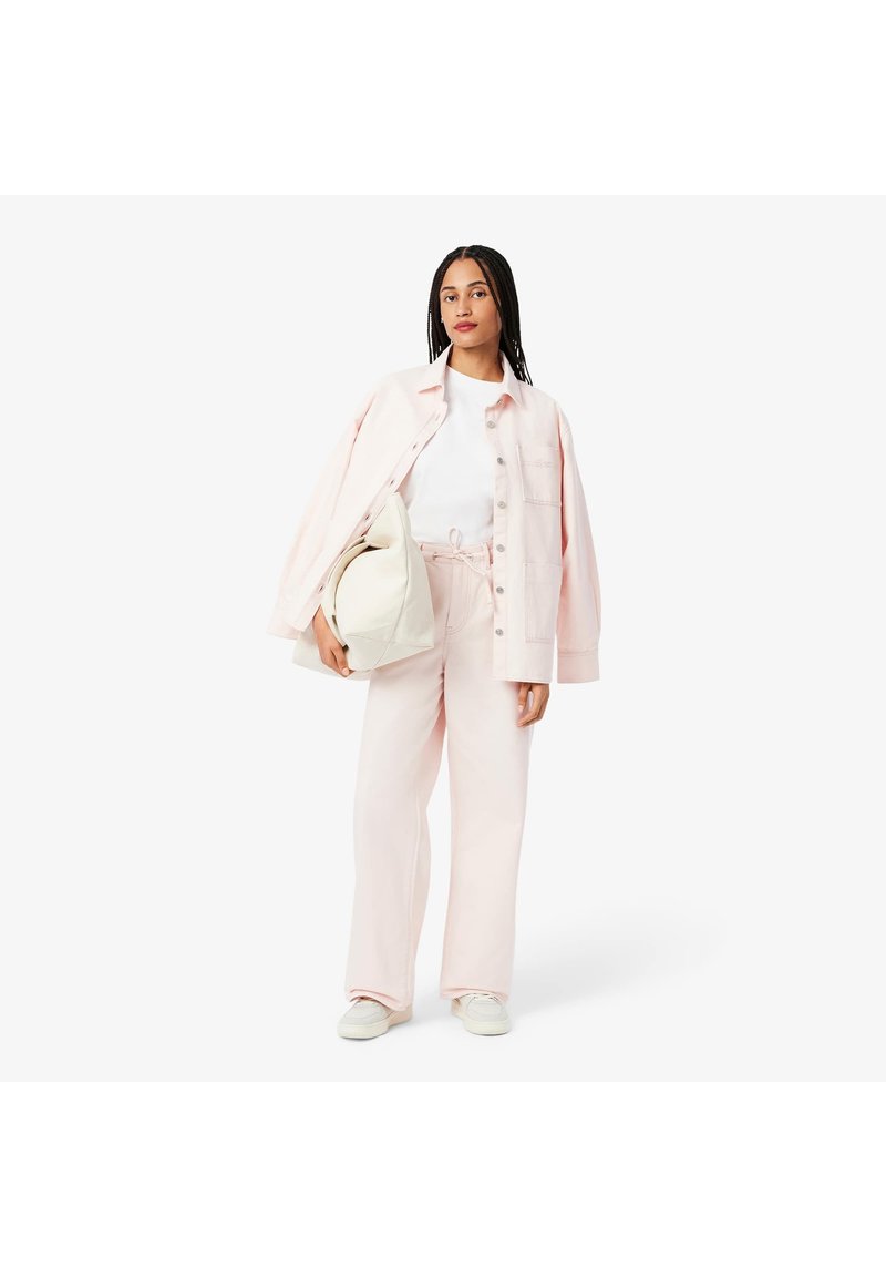 Chemise rose surdimensionnée, t-shirt blanc et pantalon rose assorti. Le modèle tient un sac crème. La tenue présente une coupe décontractée et un tissu doux.