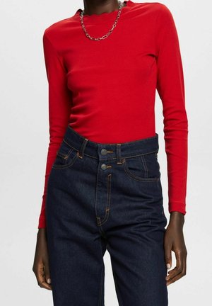 Rode lange mouwen top met een ronde hals, gecombineerd met donkerblauwe hoge taille jeans met oranje stiksels en knoopsluitingen.