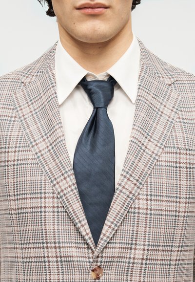 Καρό σακάκι σε καφέ και λευκό, συνοδευόμενο από σκούρα μπλε μεταξωτή γραβάτα, λευκό πουκάμισο και λεπτομέρεια με κουμπί στο γιακά.