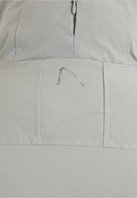 Tissu beige clair avec une fermeture éclair visible et un logo cousu ressemblant à une ligne incurvée sur un panneau segmenté.