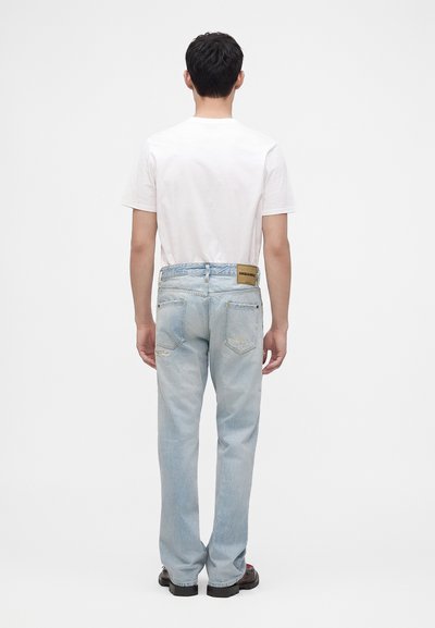 Homme de dos portant un t-shirt blanc, un jean bleu clair usé et des chaussures foncées sur un fond blanc uni.