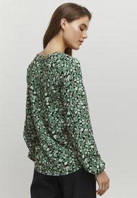 Grön blus med blommönster, lång ärm och löst snitt, med en rundad halsringning och rynkdetaljer på baksidan.