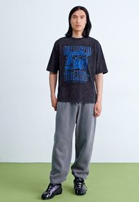 Sort grafisk t-shirt med blå tekst, parret med grå sweatpants og sorte sportssko. Glat tekstur, afslappet pasform, uformelt design.
