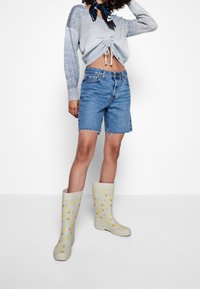 Pull court bleu clair et gris avec cordon de serrage, short en jean coupé, et bottes de pluie crème à pois multicolores.