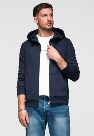 Homme portant un sweat à capuche zippé bleu marine sur un t-shirt blanc à motif graphique et un jean bleu, ajustant le col du sweat, fond clair.