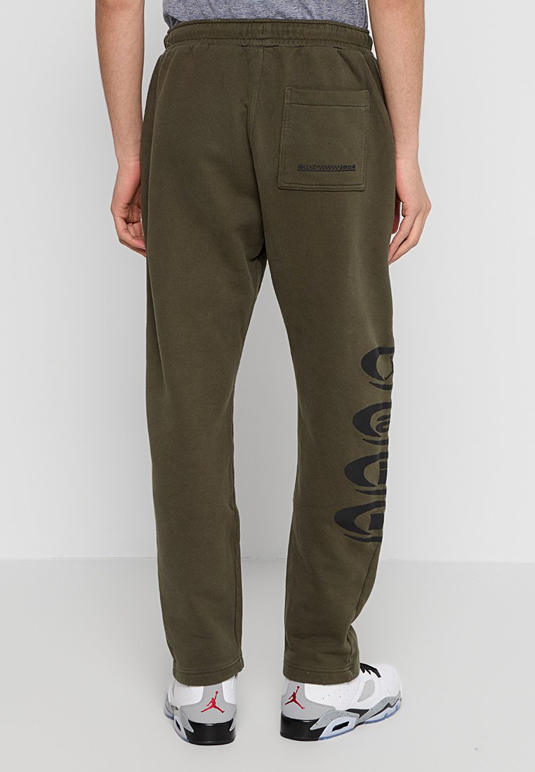 Pantalones sudadera verde oliva hechos de algodón, con un corte relajado, bolsillo lateral y un gran estampado gráfico negro a lo largo de la pierna izquierda.