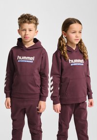 Bordowy komplet bluzy i spodni z białymi logo i paskami ozdobnymi. Materiał wydaje się miękki; projekt obejmuje kaptur i przednią kieszeń.