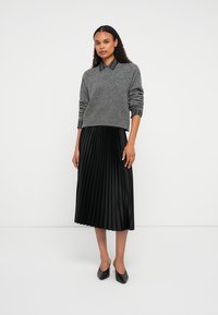 Donna in piedi che indossa un maglione grigio sopra una camicia collare nera a righe, una gonna midi plissettata nera e scarpe nere a punta.