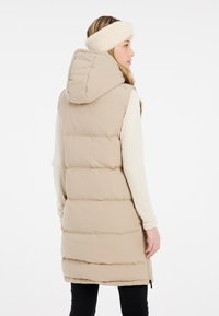 Gilet en duvet beige avec capuche, design matelassé, zips sur les côtés et silhouette longue, associé à un bandeau en polaire crème.