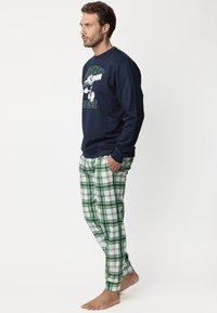 Hombre que lleva un sudadera azul marino con un estampado de Mickey Mouse y pantalones de cuadros verde y blanco, de pie descalzo con las manos en los bolsillos, mirando hacia la izquierda.