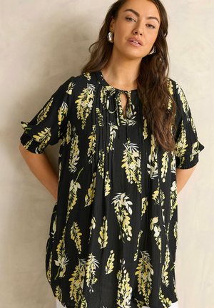 Kobieta z długimi włosami, ubrana w czarną kwiecistą bluzkę z żółtymi i białymi kwiatami, stojąca z rękami za plecami na jednolitym tle.
