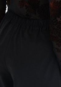 Zbliżenie czarnych spodni z elastycznym paskiem, w zestawieniu z ciemną, kwiatową wzorzystą przezroczystą bluzką z rękawami i torsu.