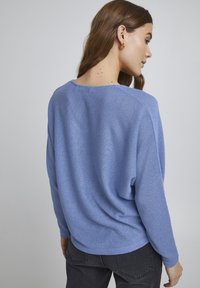Pull en tricot bleu à manches longues et coupe décontractée, avec un col large et un tissu texturé. Porté avec un jean foncé.
