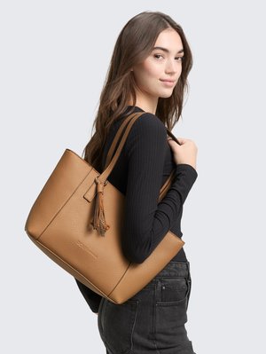 Młoda kobieta z długimi brązowymi włosami w czarnej bluzce i dżinsach, niosąca dużą jasnobrązową skórzaną torbę na ramię.