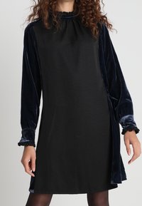 Eine Frau trägt ein schwarzes Kleid mit samtigen, langärmeligen in Marineblau und Rüschenmanschetten, steht vor einem schlichten hellen Hintergrund.