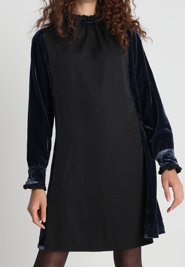 Eine Frau trägt ein schwarzes Kleid mit samtigen, langärmeligen in Marineblau und Rüschenmanschetten, steht vor einem schlichten hellen Hintergrund.