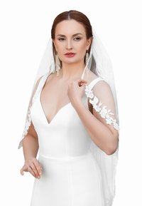 Witte trouwjurk met een diepe V-hals en bloemachtige kantmouwen. Doorzichtige sluier, opvallende oorbellen en een subtiele ring om haar vinger.