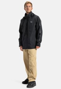 Čierna nepremokavá bunda so zipsom, nastaviteľnou kapucňou a elastickými manžetami; skombinovaná s béžovými nohavicami a čiernymi topánkami s textúrovanými detailmi.