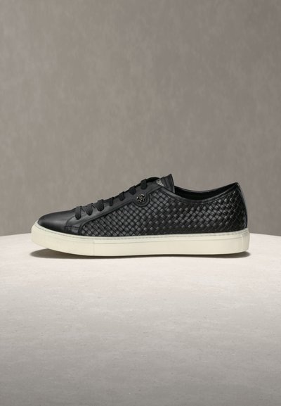 Baskets basses noires avec dessus en cuir tissé, lacets noirs et semelle en caoutchouc blanche, posées sur une surface claire avec un arrière-plan neutre.