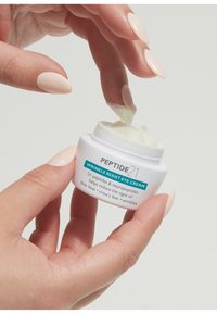 Crème contour des yeux Peptide21 Wrinkle Resist dans un pot blanc, avec une texture lisse et crémeuse. Un doigt plonge dans la crème, illustrant son application.