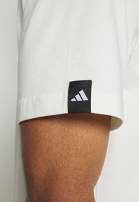 Valkoinen puuvillainen lyhythihainen paita, jossa on musta suorakulmainen merkki hihassa, jossa on kolme valkoista vinoa raitaa. Pehmeä tekstuuri, rento muotoilu.