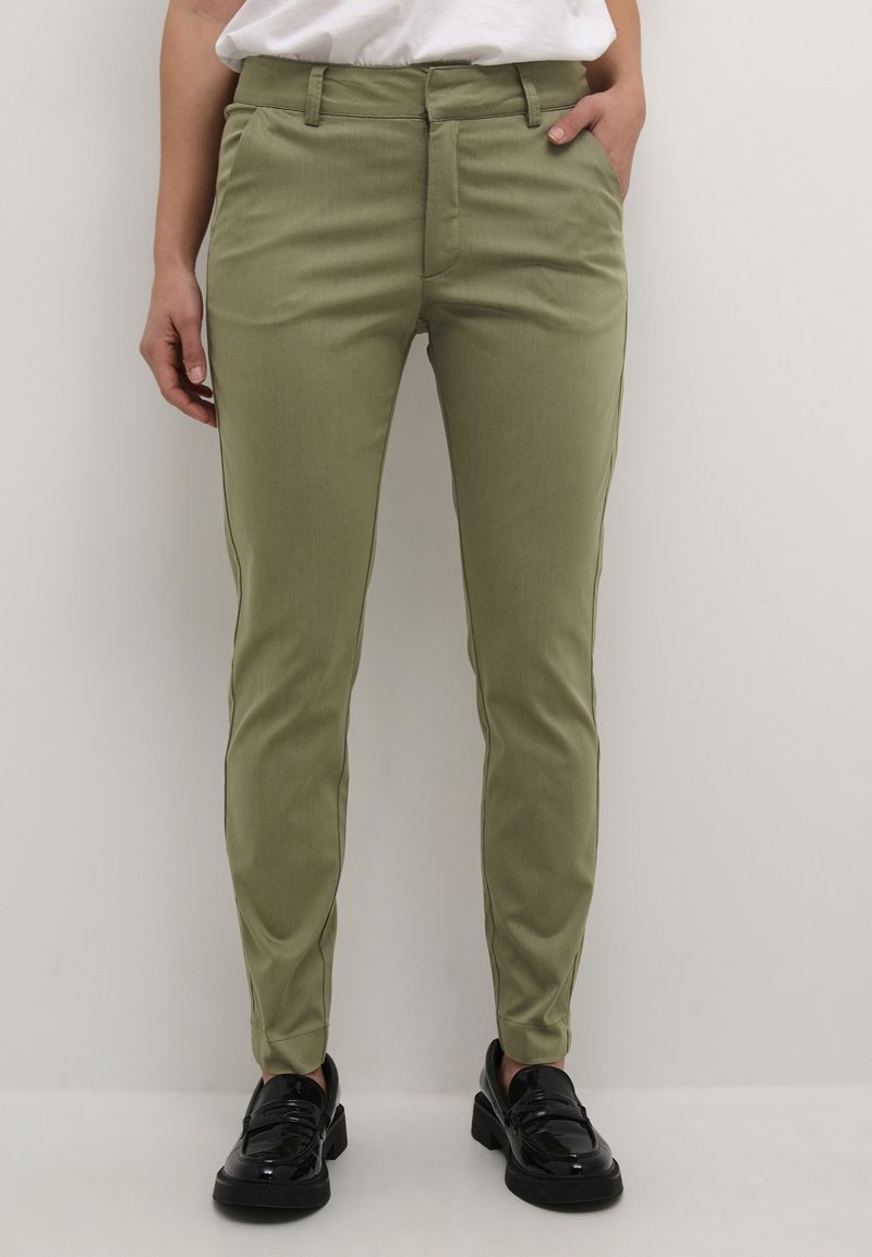 Personne portant un pantalon slim vert olive avec des poches latérales et des mocassins noirs brillants, debout devant un fond clair uni.