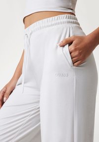 Vita sweatpants med elastisk midjeband och dragsko. Har sidofickor och en diskret "GUESS"-logga nära fickan. Mjuk textur.