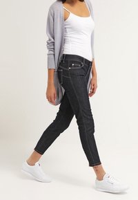 Jeans skinny de mezclilla de lavado oscuro con costuras en contraste, diseño de tiro medio y estilo de cinco bolsillos, combinados con un cardigan gris claro y zapatillas blancas.