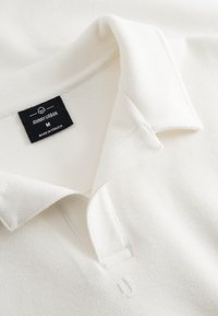 Fehér textúrájú póló ing hajtogatott gallérral, amelynek nyakkivágásában fekete címke olvasható: "JOHNNY URBAN M MADE IN TÜRKİYE".