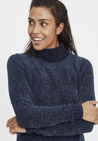 Marineblå genser med høy hals i strukturert strikkemønster, ribbet krage og mansjetter. Stoffet virker mykt og varmt, egnet for kjøligere vær.