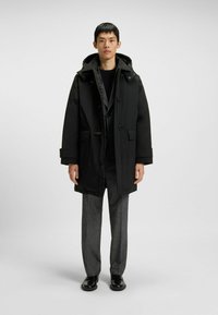 Uomo in parka nero con cappuccio sopra un completo grigio, in piedi dritto su uno sfondo grigio neutro con scarpe nere.