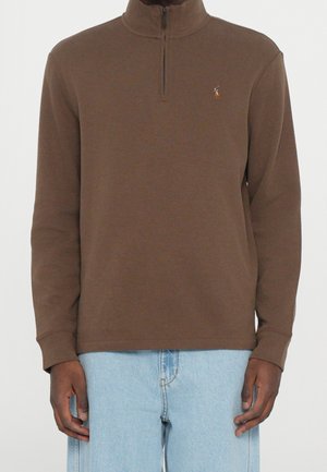 Homme portant un pull marron à manches longues avec une demi-fermeture éclair et un petit logo brodé, associé à un jean bleu clair.