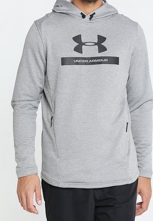 Homme portant un sweat à capuche Under Armour gris clair avec poches zippées à l'avant et pantalon noir sur fond blanc.