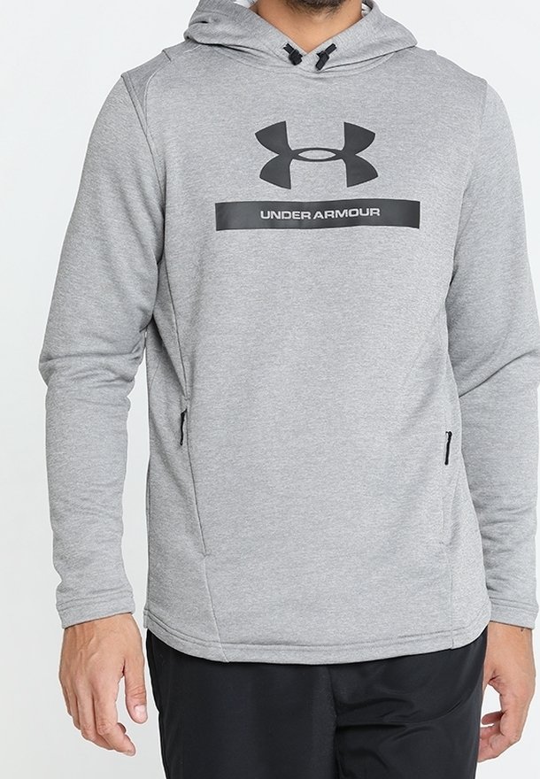 Homme portant un sweat à capuche Under Armour gris clair avec poches zippées à l'avant et pantalon noir sur fond blanc.