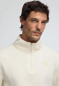 Pullover de forro polar de cremoso claro con media cremallera y un acabado texturizado, que presenta un pequeño parche de logo en relieve y un cuello alto. Se puede ver el detalle de la cremallera.