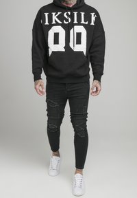 Felpa nera con stampa bianca "LIKESILF 99" e tasca frontale, abbinata a jeans skinny neri strappati e sneakers bianche. Design pulito.