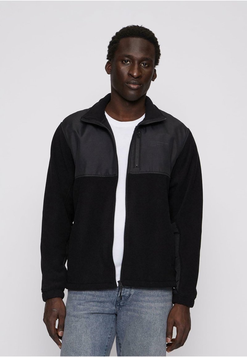 Ung mand iført sort zip-jakke over hvid skjorte og lyseblå jeans, stående mod en ensfarvet lys baggrund.