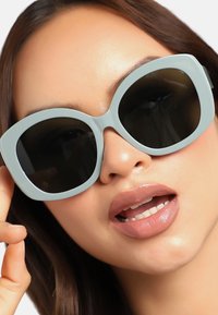 Överdimensionerade, ljusblå solglasögon med mörka linser. Tjock, glänsande båge med rundad form. Modellens läppar har en mjuk nudefärg.