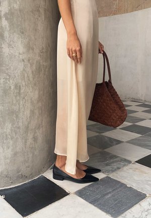 Kobieta w prześwitującej beżowej sukience, czarne niskie szpiczaste buty, trzyma brązową plecioną torebkę, stoi na czarno-białej szachownicy podłogi.