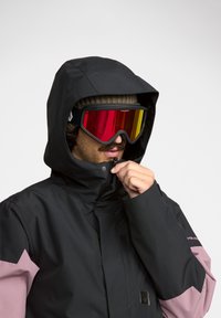 Veste isolée noire et rose avec capuche, dotée d'une fermeture éclair, d'un col ajustable et de lunettes de ski miroir avec des verres rouges et jaunes.