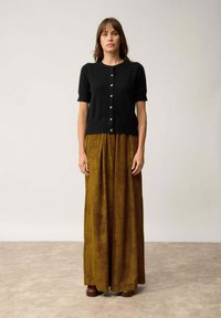 Svart kortärmad cardigan med knappar, matchad med en lång, veckad kjol i senapsgul med vertikala ränder. Bruna skor synliga.