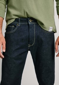 Temno modri jeans z rumenim šivom, sprednjim gumbom in petimi žepi. Kombinirani s zeleno majico z dolgimi rokavi.
